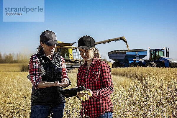 Eine reife Bäuerin steht auf einem Feld und arbeitet gemeinsam mit einer jungen Frau bei der Ernte  indem sie fortschrittliche landwirtschaftliche Software auf einem Pad verwendet  während im Hintergrund ein Mähdrescher auf einen Getreidewagen umlädt; Alcomdale  Alberta  Kanada