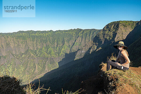 Frau mit Strohhut sitzt oben auf einer Bergklippe und blickt lächelnd in die Kamera  im Hintergrund die bergige Küste entlang des Kalalau Trail an der Napali-Küste des Pazifischen Ozeans; Kauai  Hawaii  Vereinigte Staaten von Amerika