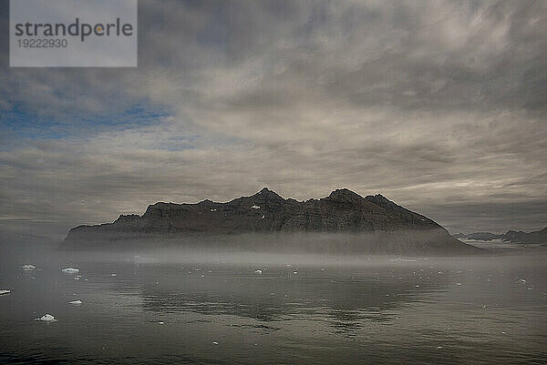 Zerklüftete Berggipfel und neblige Wolken spiegeln sich im ruhigen Wasser entlang des Nansen-Fjords; Ostgrönland  Grönland