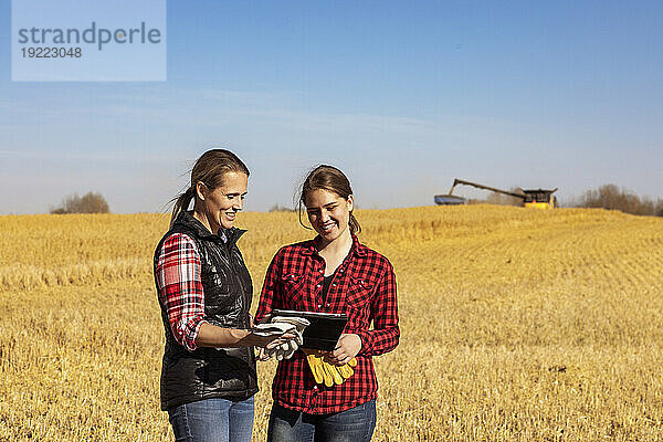 Eine reife Bäuerin  die auf einem Feld steht und ihrem Lehrling  einer jüngeren Frau  bei der Getreideernte im Herbst auf einem Pad die Verwendung fortschrittlicher landwirtschaftlicher Software beibringt  während im Hintergrund ein Mähdrescher Getreide auf einen Getreidewagen umlädt; Alcomdale  Alberta  Kanada