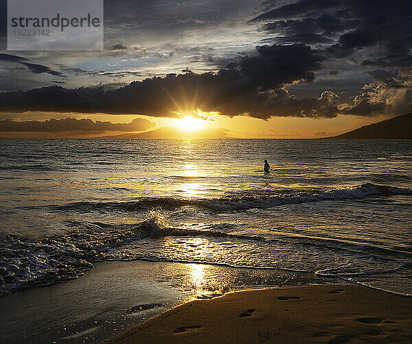 Silhouette einer Person  die in der Dämmerung im Wasser am Ufer des Strandes Kamaole 2 steht und die goldene Sonne beobachtet  die unter einem bewölkten Himmel leuchtet; Kihei  Maui  Hawaii  Vereinigte Staaten von Amerika