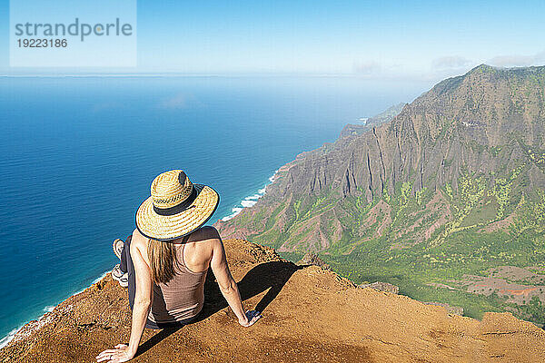 Blick von hinten auf eine Frau mit Strohhut  die auf der Spitze einer Bergklippe sitzt und auf das strahlend blaue Wasser des Pazifischen Ozeans und die bergige Küste entlang des Kalalau Trail an der Napali-Küste blickt; Kauai  Hawaii  Vereinigte Staaten von Amerika