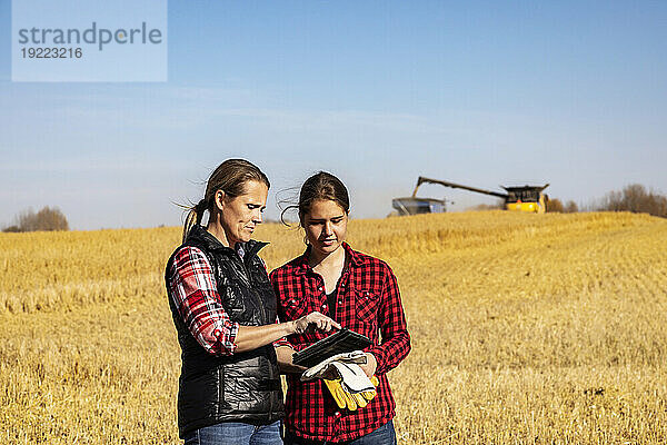 Eine reife Bäuerin  die auf einem Feld steht und ihrem Lehrling  einer jüngeren Frau  bei der Getreideernte im Herbst auf einem Pad die Verwendung fortschrittlicher landwirtschaftlicher Software beibringt  während im Hintergrund ein Mähdrescher Getreide auf einen Getreidewagen umlädt; Alcomdale  Alberta  Kanada