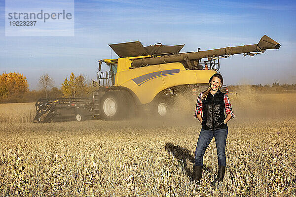 Porträt einer reifen Bäuerin  die auf einem Getreidefeld steht und während der Ernte für die Kamera posiert  während im Hintergrund bei Sonnenuntergang ein Mähdrescher arbeitet; Alcomdale  Alberta  Kanada