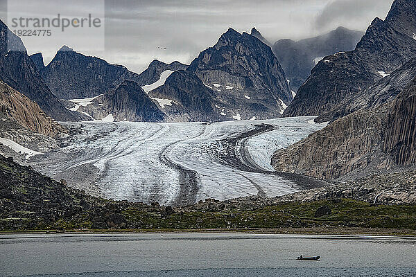 Blick auf den Gletscher  der durch die Berggipfel an der Südspitze Grönlands fließt  mit der Silhouette eines vorbeifahrenden Bootes im grauen Wasser des Prins Christian Sund; Südgrönland  Grönland