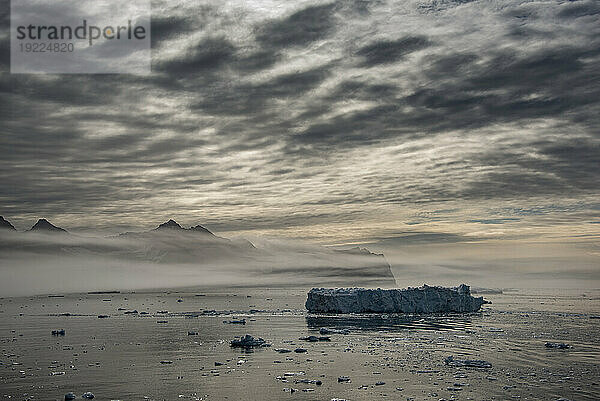 Ein Eisberg schwimmt im Gletscherwasser des Nansen-Fjords  die Silhouette der Berge zeichnet sich vor den grauen Wolken ab und ist mit einer Nebelschicht über dem ruhigen Wasser bedeckt. Ostgrönland  Grönland