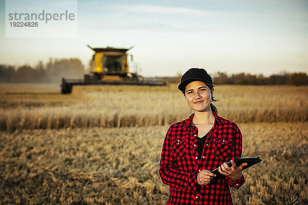 Porträt einer jungen Bäuerin  die zur Erntezeit auf einem Getreidefeld steht und fortschrittliche landwirtschaftliche Softwaretechnologien auf einem Pad nutzt  während im Hintergrund ein Mähdrescher arbeitet; Alcomdale  Alberta  Kanada