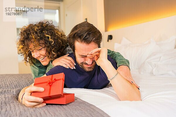 Valentinstag  ein junges kaukasisches Paar im Schlafanzug auf dem Bett  die Braut überrascht ihren Freund mit einem netten Geschenk