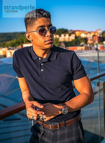 Fashion Lifestyle  Portrait eines jungen Latinos mit der Stadt San Sebastian im Hintergrund  Gipuzkoa. Blaues Polohemd und karierte Hose  nachdenklich auf der Terrasse
