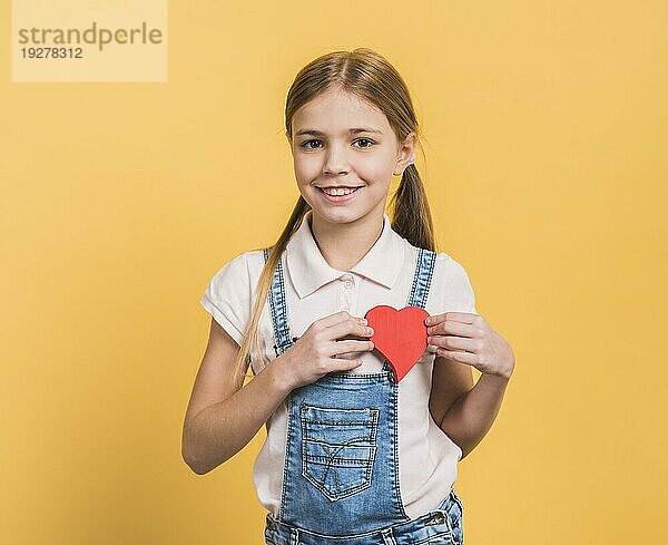 Porträt lächelndes Mädchen zeigt rote Papier ausgeschnitten Herzform gegen gelben Hintergrund