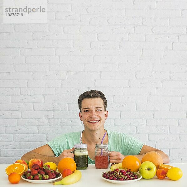 Glücklicher junger Mann hält Smoothies mit bunten frischen Biofrüchten in der Hand