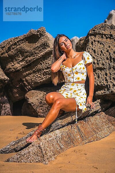 Ein brünettes Mädchen in einem weißen Kleid am Strand genießen den Sommer  sitzen auf einigen Felsen