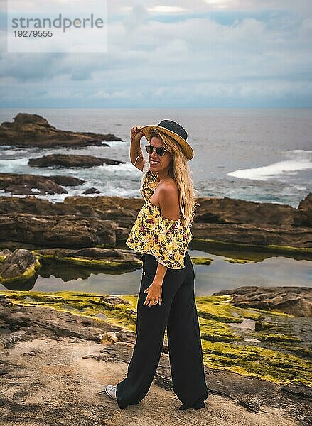 Kaukasisches blondes Mädchen in einem geblümten Hemd  schwarzen Shorts und Strohhut in einer natürlichen Landschaft am Meer und Felsen bei Sonnenuntergang  Lifestyle. Posing stehend am Meer