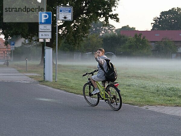 Kind  Junge mit Fahrrad und Schultasche  winkt  am Morgen am Wiesenrand mit Nebel  Schlepzig  Brandenburg  Deutschland  Europa