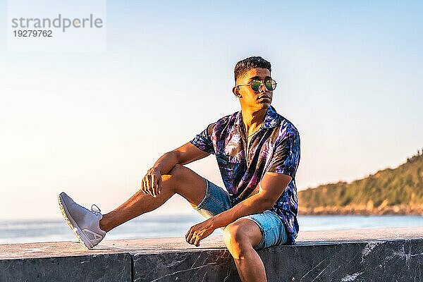 Sommerlicher Lebensstil  Porträt eines jungen Latinomannes der den Sommerurlaub genießt. mit aufgeknöpftem lila Blumenhemd und Sonnenbrille