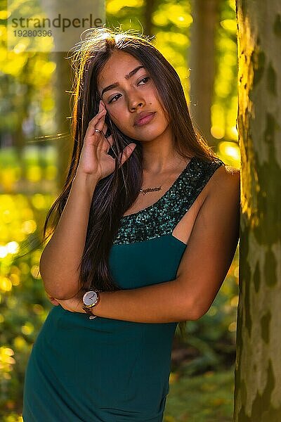 Eine junge brünette Latina mit langen glatten Haaren lehnt in einem grünen Kleid an einem Baum. Porträt mit einem süßen und sinnlichen Blick  vertikales Foto