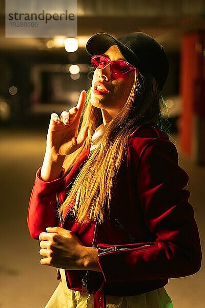 Fotografie mit roten und grünen Neons auf einem Parkplatz. Junge hübsche blonde kaukasische Frau in einem roten Anzug  Sonnenbrille und einer schwarzen Kappe