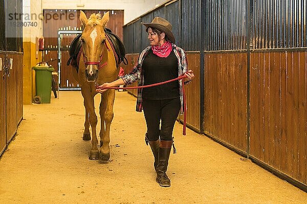 Kaukasische erwachsene Frau in einem Pferdestall  die mit einem braunen Pferd spazieren geht  gekleidet in südamerikanischer Kleidung  arbeitet mit Pferden