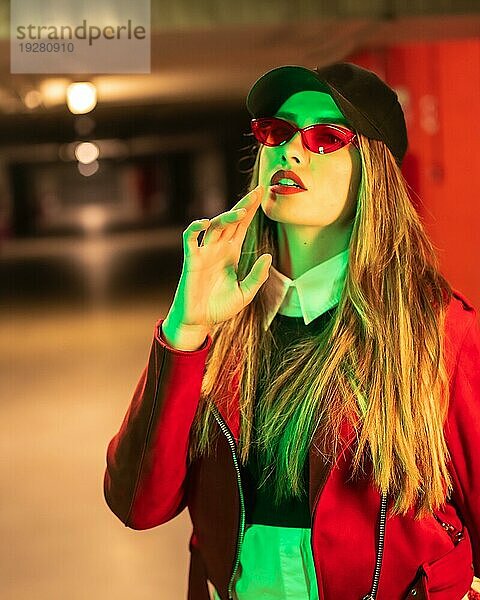 Fotografie mit roten und grünen Neons auf einem Parkplatz. Junge hübsche blonde kaukasische Frau in einem roten Anzug  Sonnenbrille und einer schwarzen Kappe