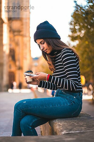 Lifestyle  brünette Kaukasierin  die eine Nachricht mit ihrem Handy in der Stadt sendet. Entspannt sich mit einem Kaffee zum Mitnehmen neben einer schönen Kirche im Park. Schwarzer Pullover und eine Wollmütze