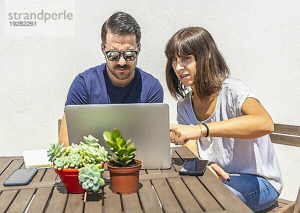 Ein Ehepaar sitzt zu Hause auf der Terrasse seines Hauses vor einem weißen Hintergrund an einem Tisch und führt ein Videogespräch mit einigen Freunden am Computer. Zeigen Sie auf eine Aktion in dem Videoanruf