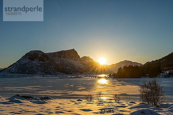 Sonnenuntergang hinter schneebedeckten Bergen über zugefrorenem See in verträumter Winterlandschaft auf den Lofoten in Norwegen an einem klaren Wintertag