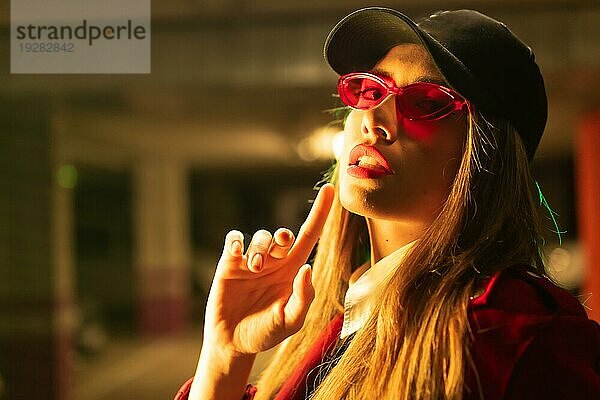 Fotografie mit roten und grünen Neons auf einem Parkplatz. Porträt einer jungen blonden kaukasischen Frau in einem roten Anzug  Sonnenbrille und einer schwarzen Kappe