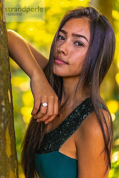 Eine junge hübsche brünette Latina mit langen glatten Haaren lehnt in einem grünen Kleid an einem Baum. Porträt von mit süßer Mida im Park