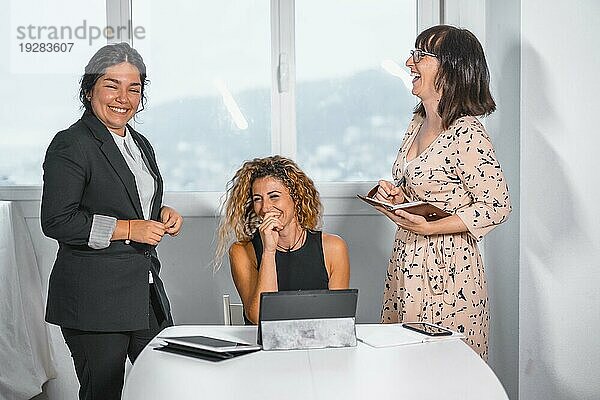 Junge unternehmungslustige Sitzung im Büro  zwei junge kaukasische Mädchen und eine junge Latina im Büro unterhalten sich fröhlich miteinander