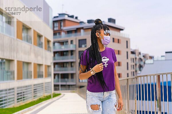 Internationaler Frauentag im Jahr der Coronaviruspandemie Covid 19. Dominikanische Frau mit Zöpfen und feministischem lila Hemd und Gesichtsmaske in der Stadt