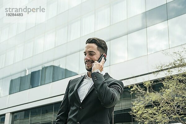 Lächelnder junger Geschäftsmann  der vor einem Bürogebäude steht und mit einem Mobiltelefon spricht