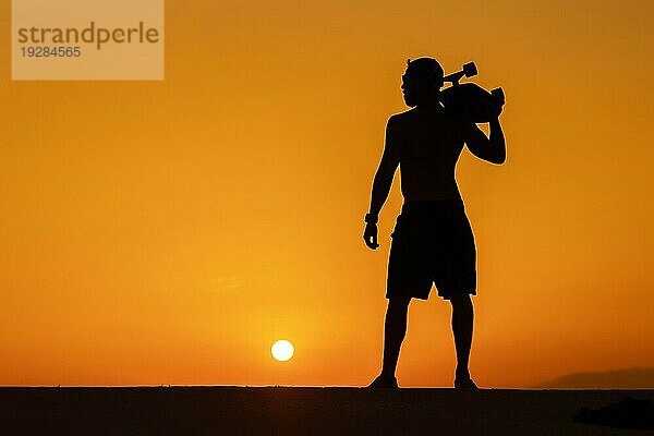 Silhouette eines freien Mannes  der bei hellem Sonnenuntergang ein Skateboard auf der Schulter trägt. Mittlere Einstellung