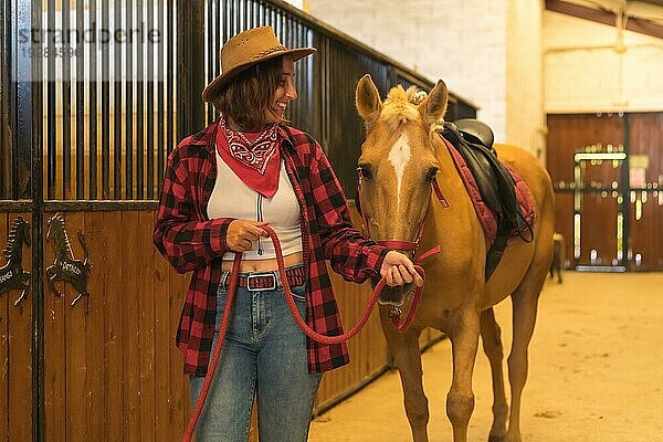 Kaukasische Cowgirl Frau  die sich mit einem Pferd in einem Stall amüsiert und lächelt  amerikanischer Cowboyhut  rotkariertes Hemd und Jeans
