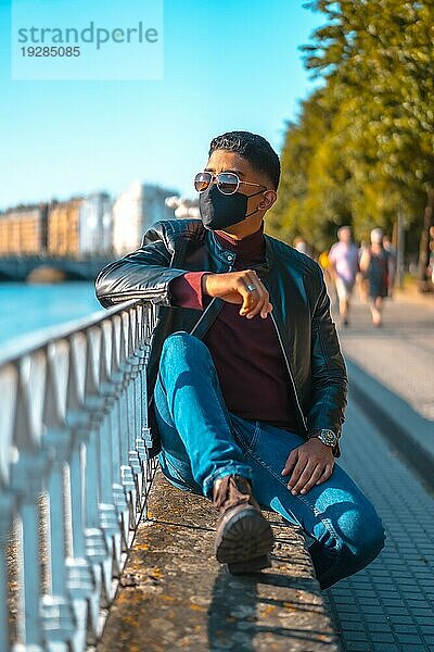Mode Lebensstil  Porträt eines jungen Latino in den Fluss der Stadt. Jeans  Lederjacke und braune Schuhe. In einer Pandemie mit einer Maske