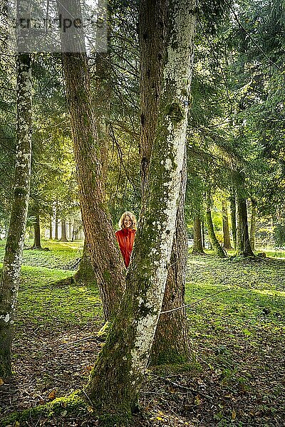 Eine Frau steht in einer Parklandschaft bzw. Waldlandschaft und schaut lachend zwischen Baumstämmen hindurch. Die herbstliche Morgensonne scheint zwischen den Bäumen hindurch. Isny im Allgäu  Deutschland  Europa