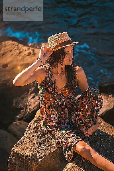 Eine junge kaukasische Frau mit nassen Haaren  einem Blumenkleid und einem Hut  die auf einem Felsen am Meer sitzt  Sommerleben in der goldenen Stunde