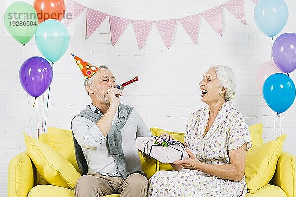 Glückliche Frau mit Geburtstagsgeschenk  die ihren Mann beim Blasen des Partyhorns sieht