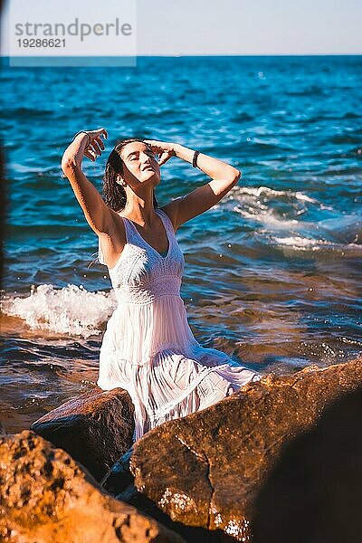 Ein junges kaukasisches Mädchen lächelnd in einem nassen weißen Kleid auf einem Felsen am Meer sitzend  Sommerleben