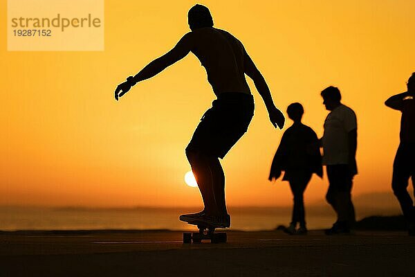 Silhouette eines Mannes  der bei Sonnenuntergang um andere Menschen herum Skateboard fährt. Mittlere Aufnahme