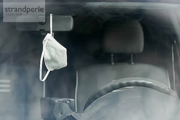 Weiße Schutzmaske  die am Rückspiegel im Auto hängt  von vorne gesehen durch die Frontscheibe gesehen