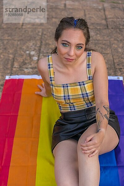 Vertikale Aufnahme einer jungen lächelnden kaukasischen Frau  die auf einer LGBT Flagge im Freien sitzt