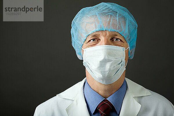 Männlicher Arzt mit Maske und Mütze schaut in die Kamera