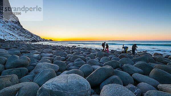 Gruppe von Landschaftsfotografen am berühmten Strand bei Uttakleiv mit runden Felsblöcken auf den Lofoten in Norwegen an einem klaren Wintertag mit schneebedeckten Bergen und blauem Himmel nach Sonnenuntergang