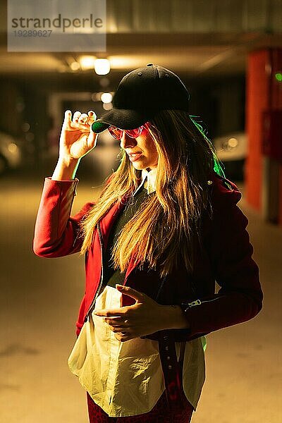 Fotografie mit roten und grünen Neons auf einem Parkplatz. Junge hübsche blonde kaukasische Frau in einem roten Anzug  Sonnenbrille und einer schwarzen Kappe