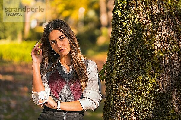 Eine junge kaukasische Brünette  die an einem Herbstnachmittag in einem Park neben einem Baum sitzt