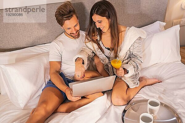 Ein Paar im Schlafanzug auf der Suche nach Hotels oder Ausflügen vom Computer aus beim Frühstück im Hotelbett  Lifestyle eines verliebten Paares