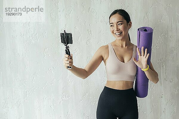 Smileyfrau die mit ihrem Telefon einen Vlog führt  während sie eine Fitnessmatte hält