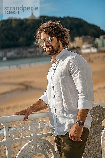 Sommerlicher Lebensstil mit einem jungen dunkelhaarigen kaukasischen Mann mit langem Bart  Sonnenbrille und weißem Hemd an einem Strand. San Sebastian  Baskenland. Genießen Sie den Strand im Sommer