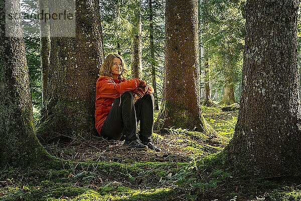 Eine Frau sitzt entspannt in einer Parklandschaft bzw. Waldlandschaft  lehnt an einem Baum und genießt die Natur beim Waldbaden. Die herbstliche Morgensonne scheint zwischen den Bäumen hindurch. Isny im Allgäu  Deutschland  Europa