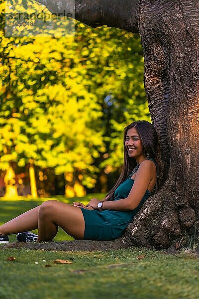 Eine junge hübsche Latina Brünette mit langen glatten Haaren lehnt in einem grünen Kleid an einem Baum. Sitzen an einem Baum im Park